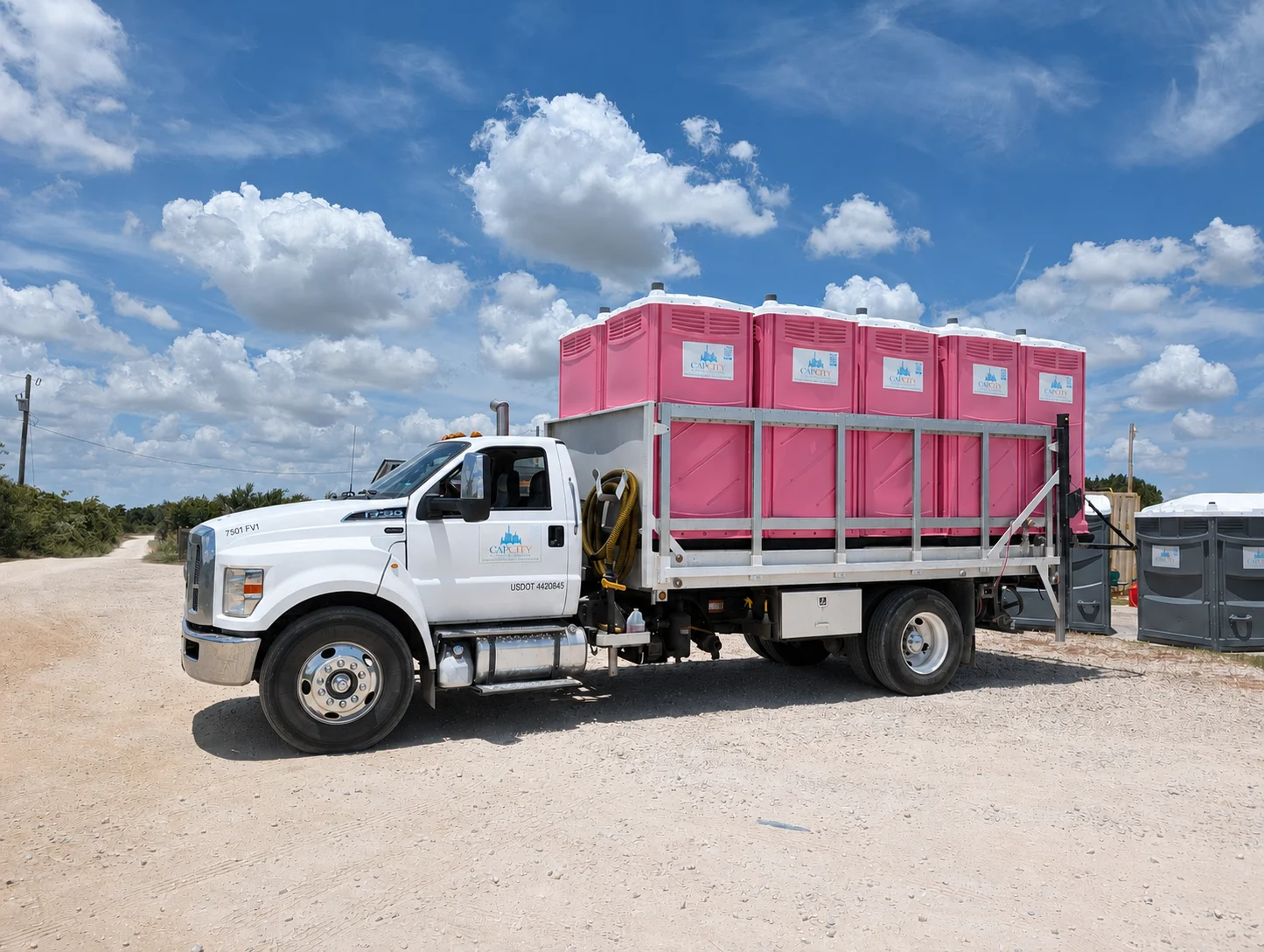 A white Ford F-750 flatbed truck loaded with five pink portable toilets branded "Cap City," parked on a dirt lot under a blue sky with scattered white clouds.